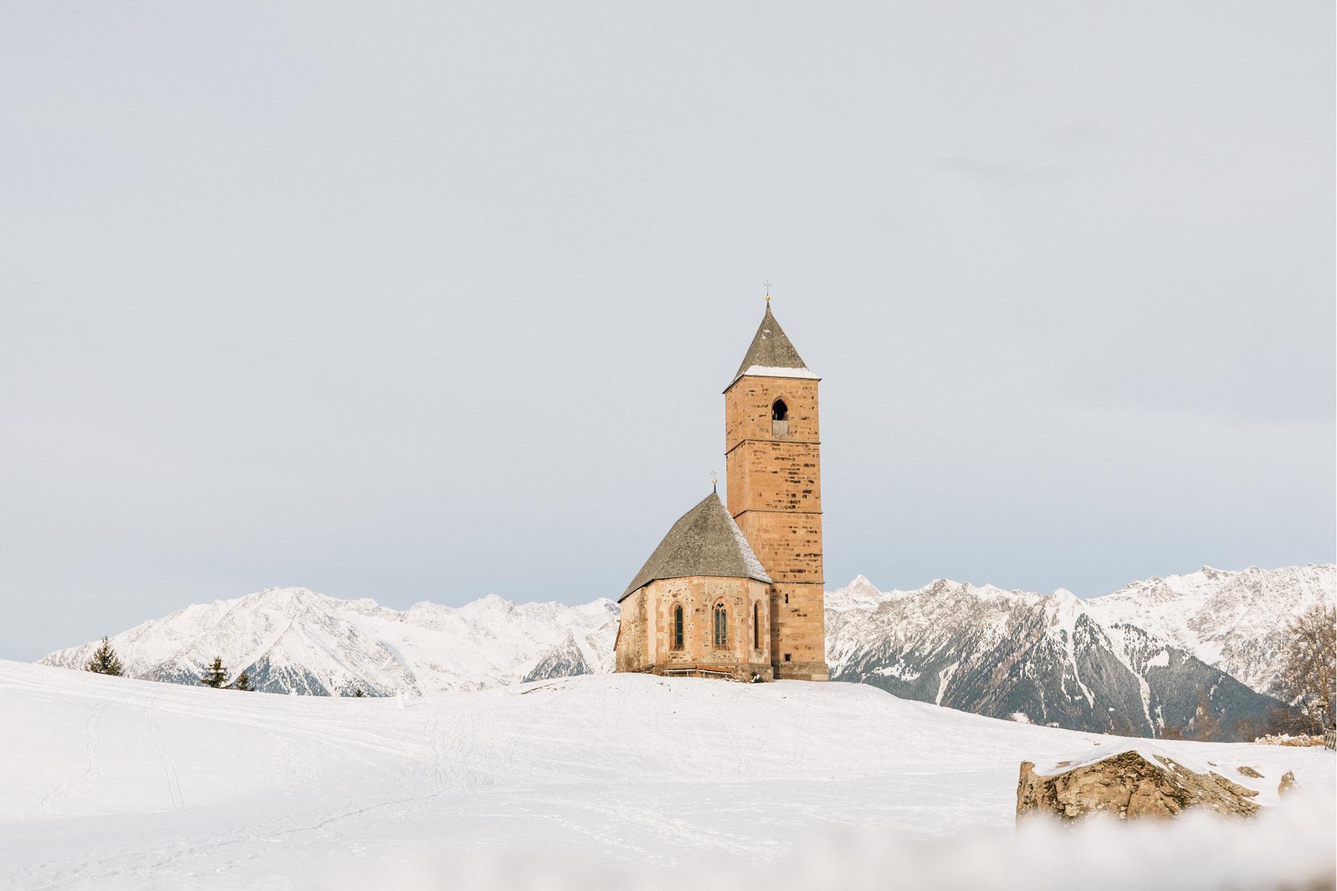 Die St. Kathrein Kirche in der Scharte in Hafling umgeben von frischem pulverschnee. Im Hintergrund die weißen Bergspitzen im Winter. Bisschen vor der Kirche der markante Stein.