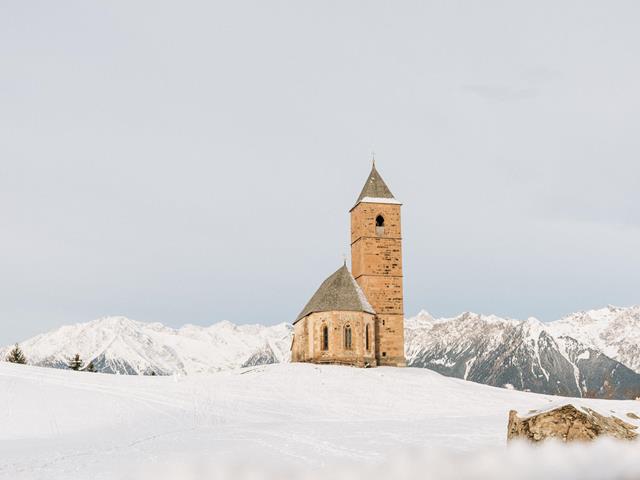 Die St. Kathrein Kirche in der Scharte in Hafling umgeben von frischem pulverschnee. Im Hintergrund die weißen Bergspitzen im Winter. Bisschen vor der Kirche der markante Stein.