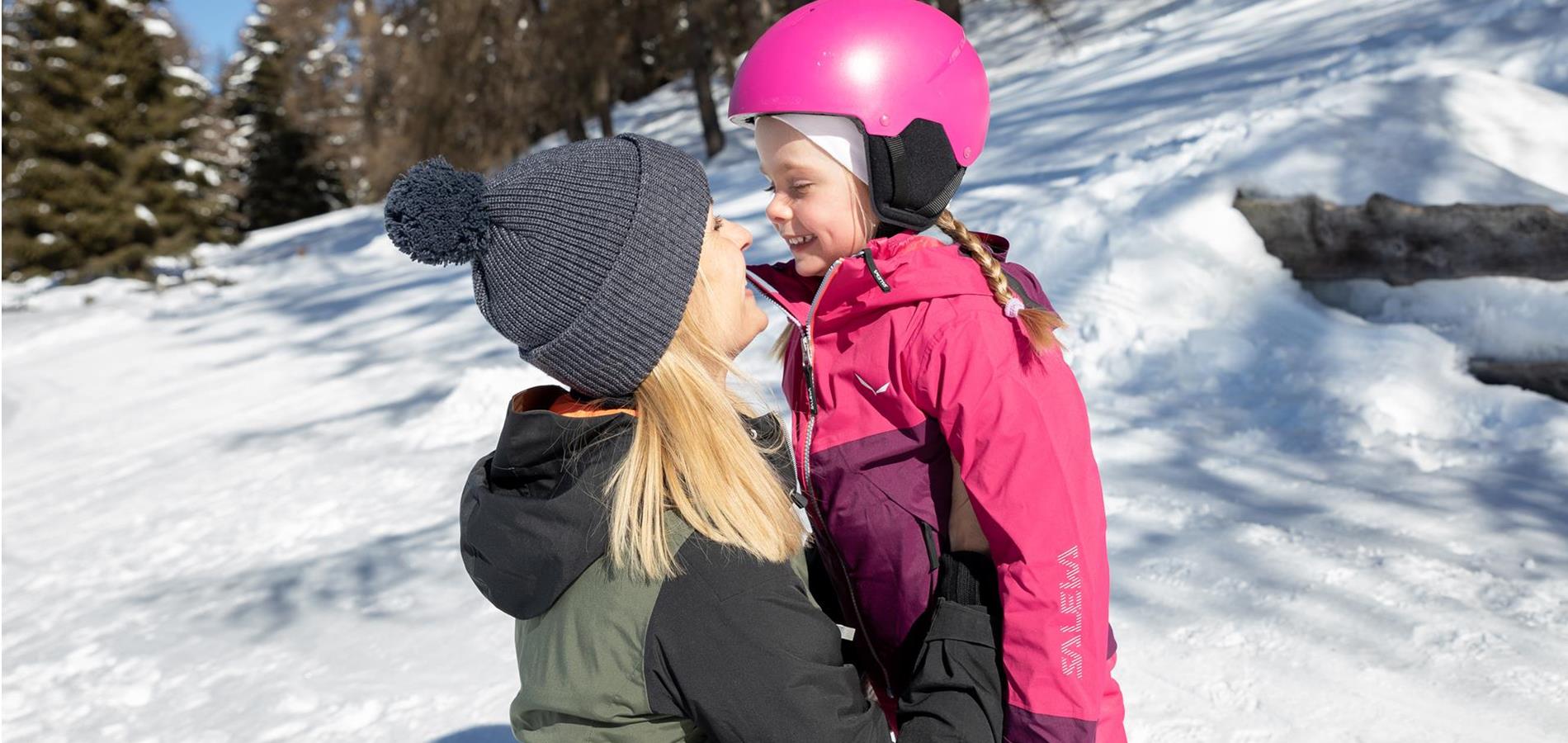 Mother and daughter in winter in Verano, South Tyrol. Both are laughing and looking at each other. The girl is around 4 years old and is wearing a pink ski suit with a pink helmet.